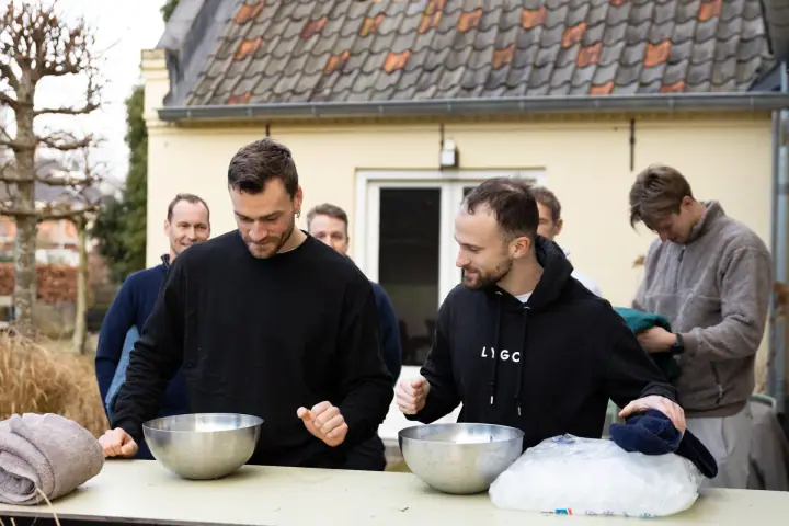 Twee mannen zetten zich lachend schrap boven hun schaal ijswater, naast een zak ijsblokjes