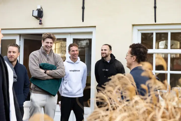 Vier deelnemers glimlachen naar de coach, die half van de camera afgedraaid achter het siergras staat