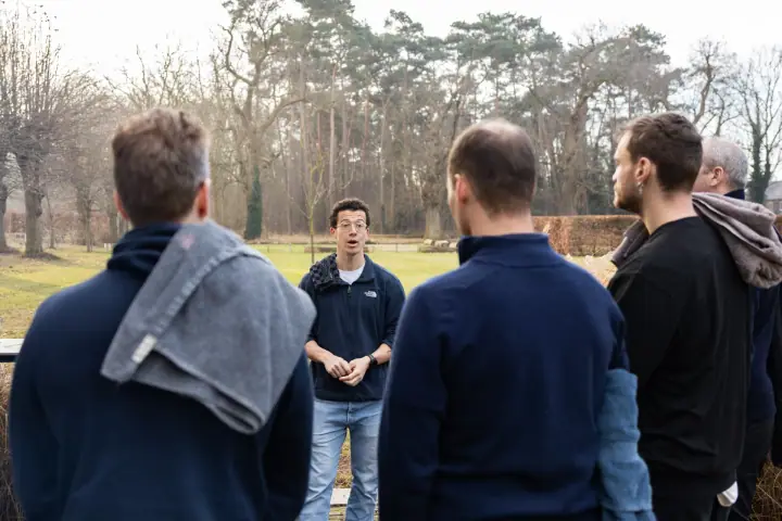 Deelnemers met een handdoek over de schouder luisteren in de tuin naar de uitleg van de coach