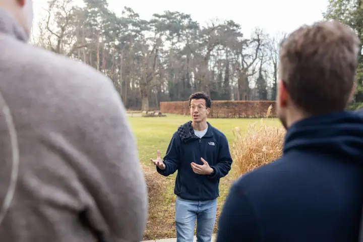 Met open handen legt de coach iets uit aan de groep, met het gazon en een beukenhaag achter zich