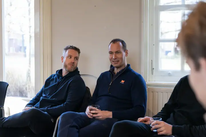 Twee deelnemers zitten binnen bij het raam met een glas in de hand en luisteren aandachtig