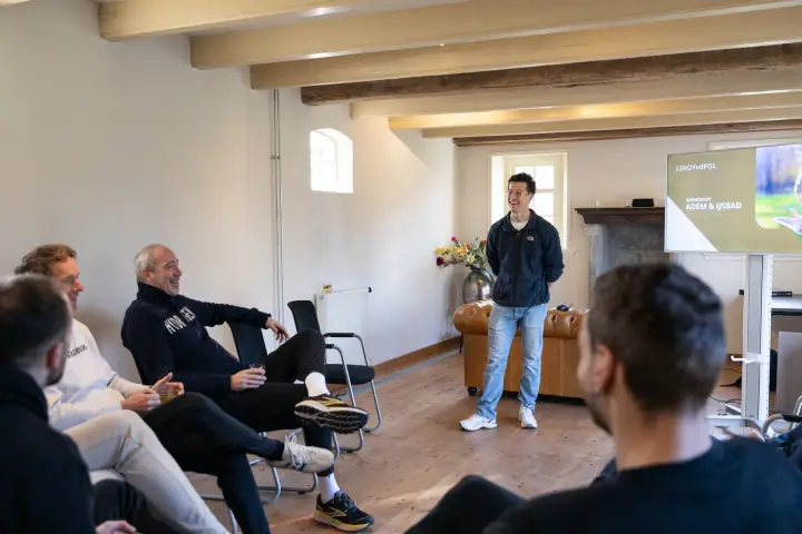 De coach lacht voor de groep in een zaal met houten balken, op het scherm staat workshop adem en ijsbad
