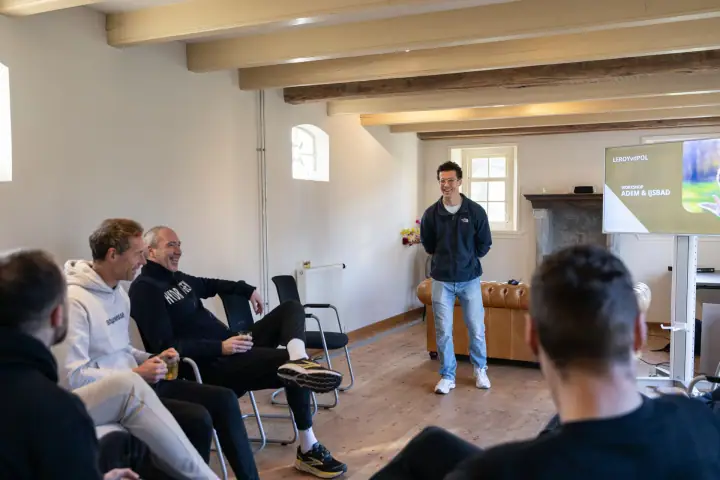 Deelnemers zitten in een kring op stoelen en lachen mee met de coach tijdens het theoriedeel
