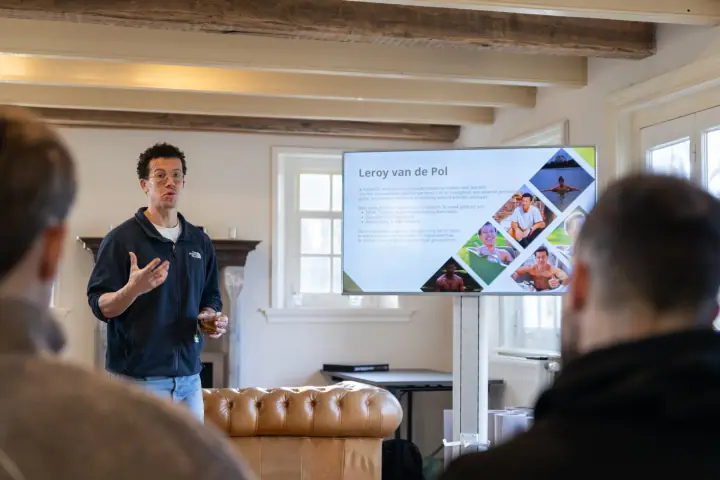 De coach presenteert naast een scherm met slides over zijn achtergrond, voor een leren chesterfieldbank