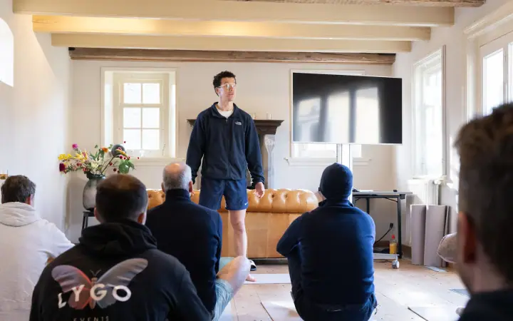 De coach in korte broek staat voor de groep, die op matjes op de houten vloer zit