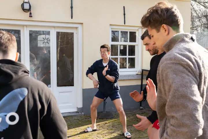 Buiten doet de coach een houding voor en de deelnemers bootsen hem na met geheven handen