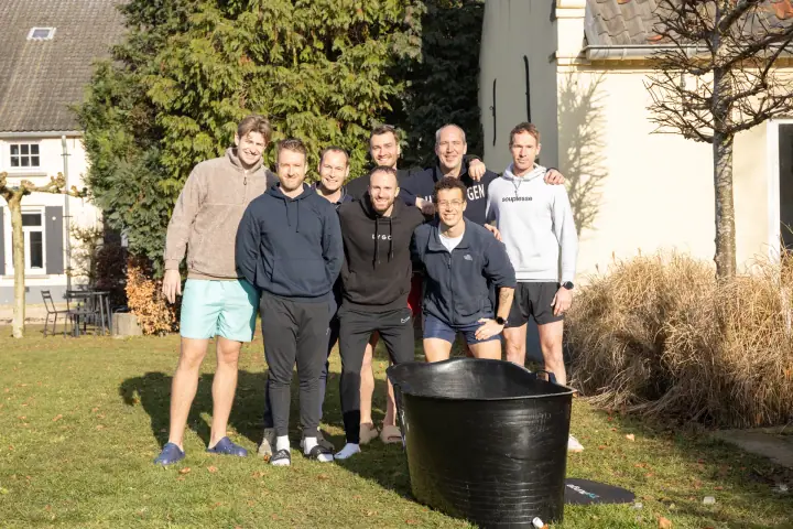 De deelnemers en de coach poseren samen op het gazon, met de zwarte ijsbadkuip vooraan