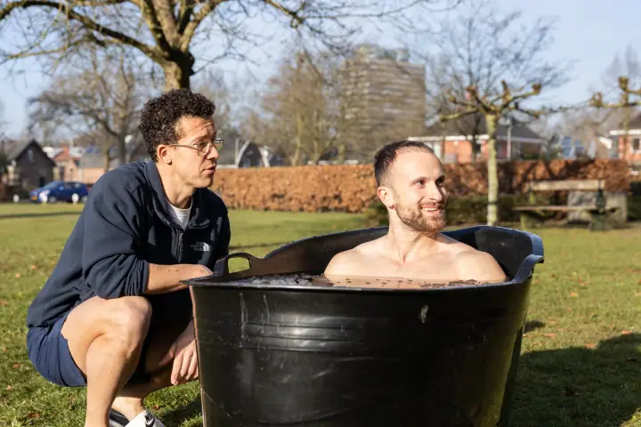 De coach zit gehurkt naast een glimlachende deelnemer die tot aan zijn schouders in het ijsbad zit