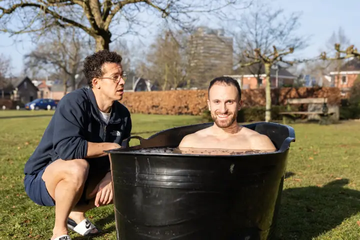 Een deelnemer kijkt glimlachend in de camera vanuit de ijsbadkuip, terwijl de coach hem aandachtig in de gaten houdt