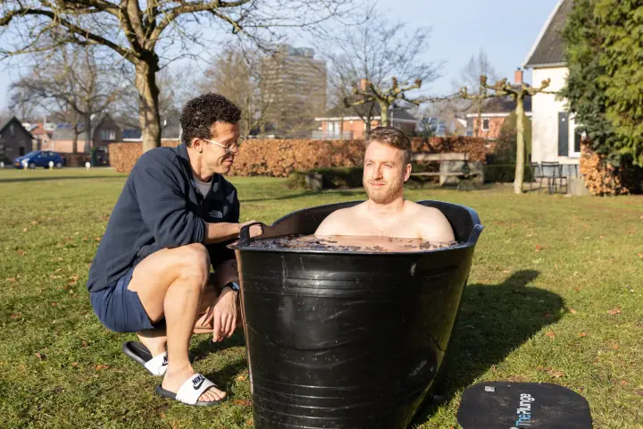 Een deelnemer zakt in de zwarte ijsbadkuip op het gras terwijl de coach hem rustig begeleidt