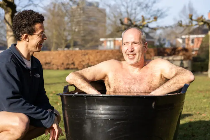 Lachende deelnemer met zijn armen over de rand van het ijsbad, de coach lacht met hem mee