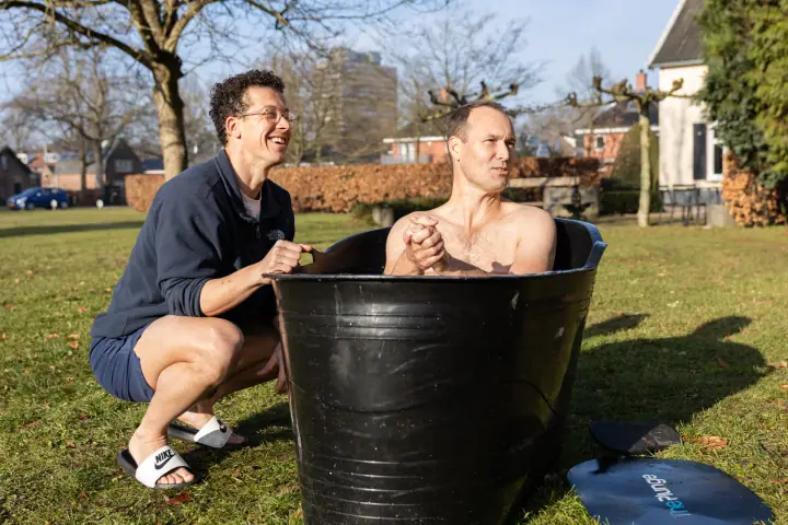 De coach lacht terwijl een deelnemer met gevouwen handen in de ijsbadkuip zit, op een zonnige winterdag