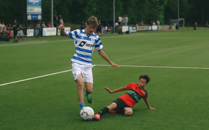 NEC-speler gaat er met een sliding in, de jeugdspeler van De Graafschap stapt over zijn been heen