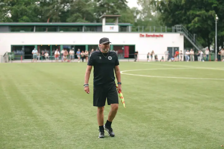 Grensrechter loopt over het veld met het clubgebouw van De Eendracht en toeschouwers op de achtergrond