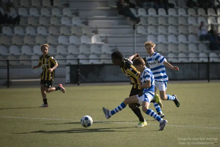 Speler van De Graafschap steekt zijn been uit naar de bal van een Vitesse-speler, lege stoeltjes erachter