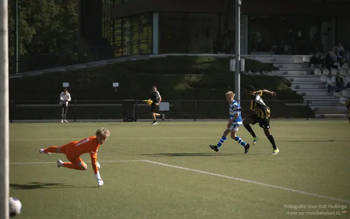 Keeper in oranje strekt zich naar de bal bij de paal, twee spelers lopen in de verte mee