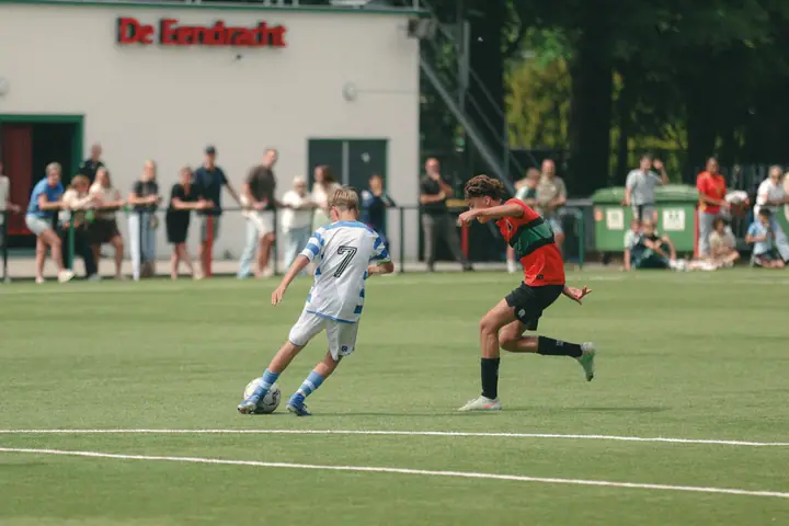 Nummer 7 van De Graafschap neemt de bal mee terwijl een NEC-speler aanstormt, publiek achter het hek