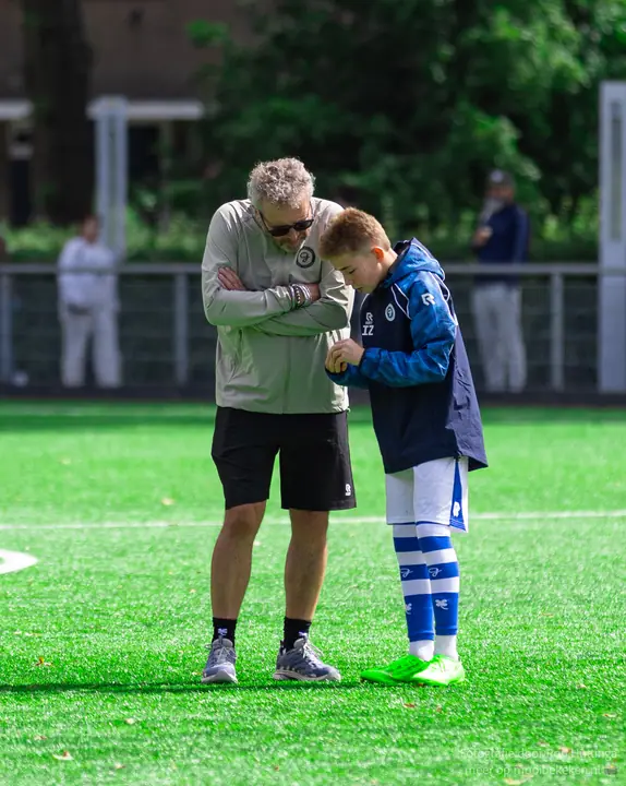 Staflid met de armen over elkaar bespreekt iets met een jeugdspeler in trainingsjack