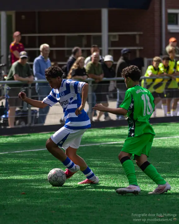 Speler van De Graafschap draait met de bal weg van nummer 10 in het groen, toeschouwers achter het hek