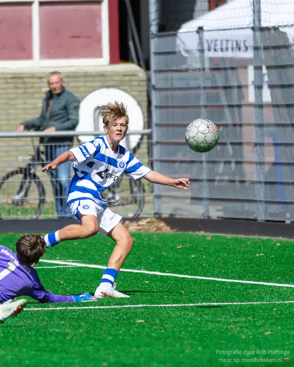 De bal vliegt hoog weg terwijl de keeper in paars op de grond ligt en de schutter hem nakijkt