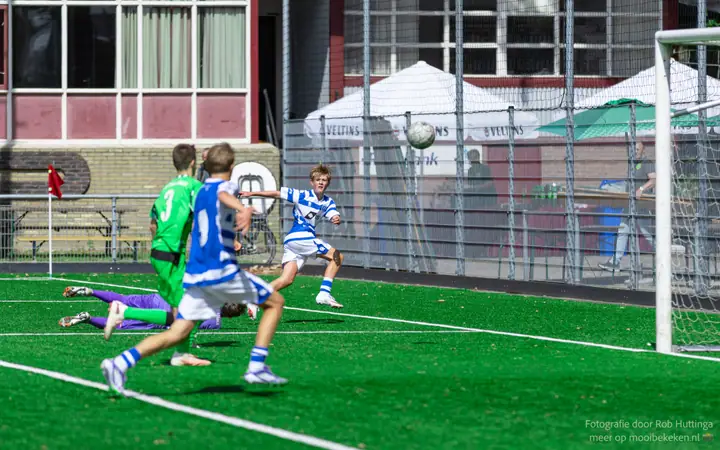 Bal zweeft richting het doel terwijl de keeper in paars gevloerd ligt en spelers toekijken
