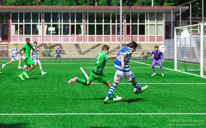 Speler van De Graafschap haalt uit terwijl nummer 3 in het groen probeert te blokken, de keeper wacht
