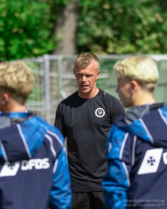 Trainer in zwart shirt spreekt zijn jeugdspelers in blauwe trainingsjacks toe