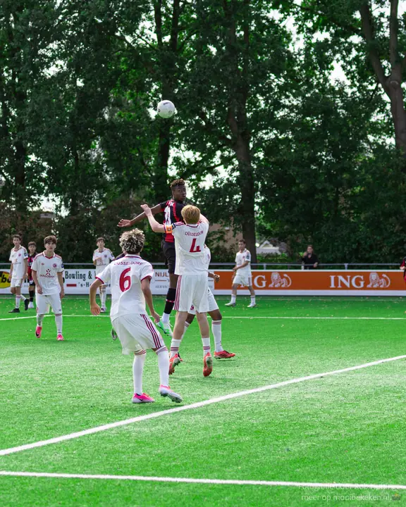 Kopduel tussen een speler in rood-zwart en twee verdedigers in wit-rood tenue voor een oranje reclamebord