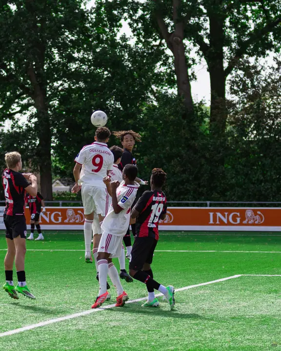 Nummer 9 in het wit wint de kopbal boven spelers in rood-zwarte shirts