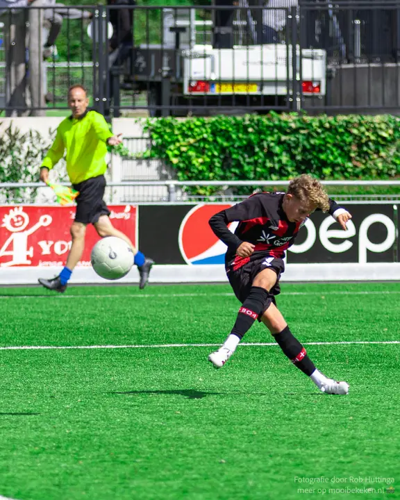 Speler in zwart-rood tenue schiet op doel, met de grensrechter in geel op de achtergrond