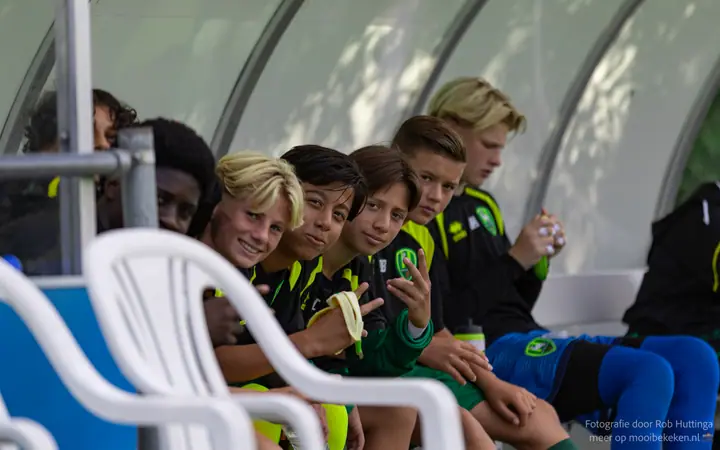 Wisselspelers in zwart-geel trainingstenue zitten lachend op de bank in de dug-out