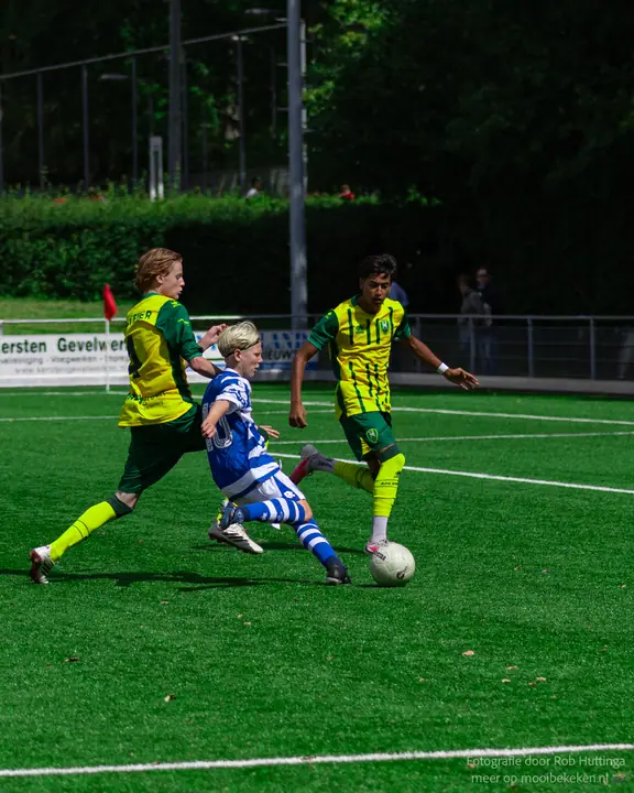 Jeugdspeler van De Graafschap glijdt tussen twee tegenstanders in geel-groen naar de bal