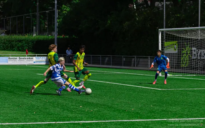 Blonde speler van De Graafschap prikt de bal weg tussen twee tegenstanders in geel-groen, de keeper in blauw wacht