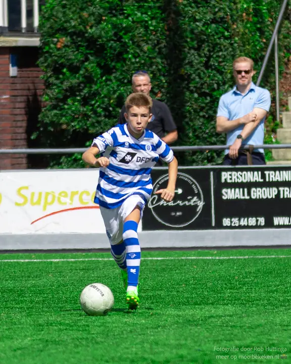 Jeugdspeler van De Graafschap loopt met de bal recht op de camera af, toeschouwers achter de boarding