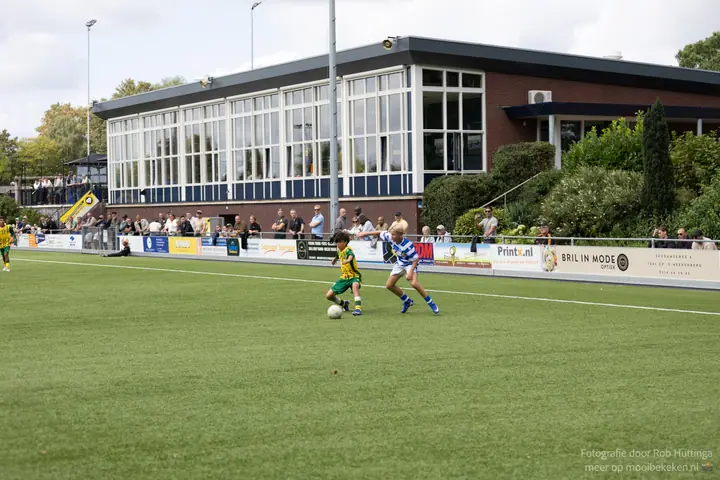Breed beeld van het sportpark met clubgebouw, waar een speler van De Graafschap en een tegenstander strijden om de bal