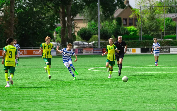 Jeugdspeler van De Graafschap laat de bal lopen tussen spelers in geel-groen, de scheidsrechter staat ernaast