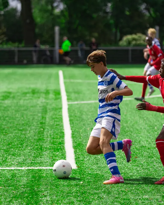 Jeugdspeler van De Graafschap neemt de bal mee langs de lijn, een tegenstander in rood duwt in zijn rug