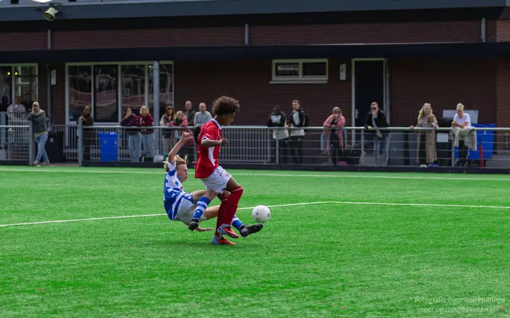 Speler van De Graafschap gaat liggend in de tackle bij een tegenstander in rood, voor het clubgebouw
