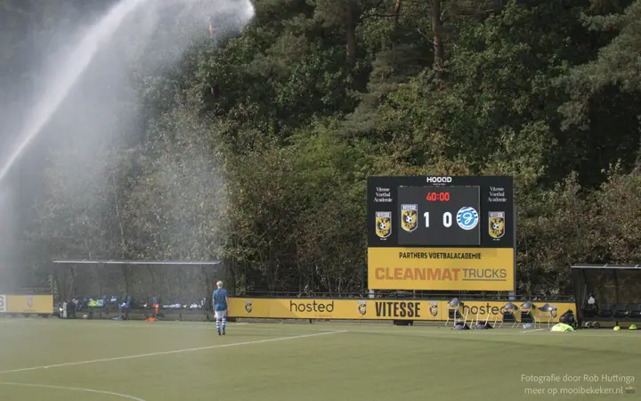 Sproeiers zetten het veld bij Vitesse onder water terwijl het scorebord een 1-0 tussenstand toont