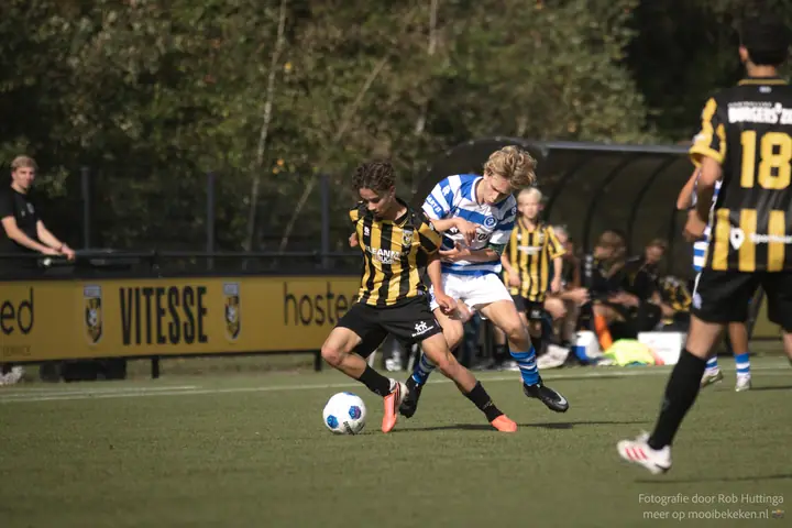 Fysiek duel om de bal met een Vitesse-speler, voor de reclameborden langs het veld