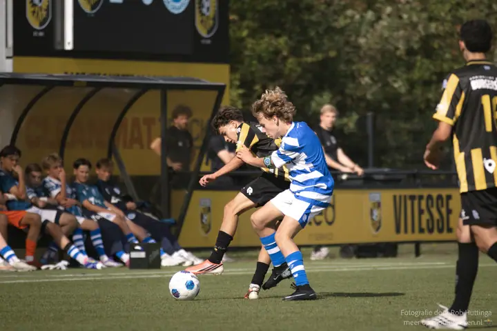 Duel tussen een speler van Vitesse en De Graafschap, met de wisselspelers op de bank erachter
