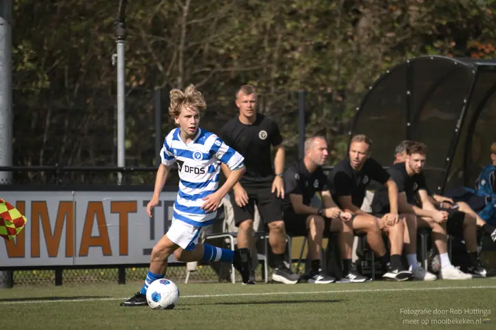 Jeugdspeler van De Graafschap dribbelt langs de lijn, met de staf in het zwart op de bank erachter