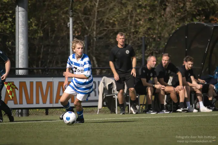 Blonde speler van De Graafschap kijkt op voor een pass, een staflid staat vlak achter hem langs de lijn