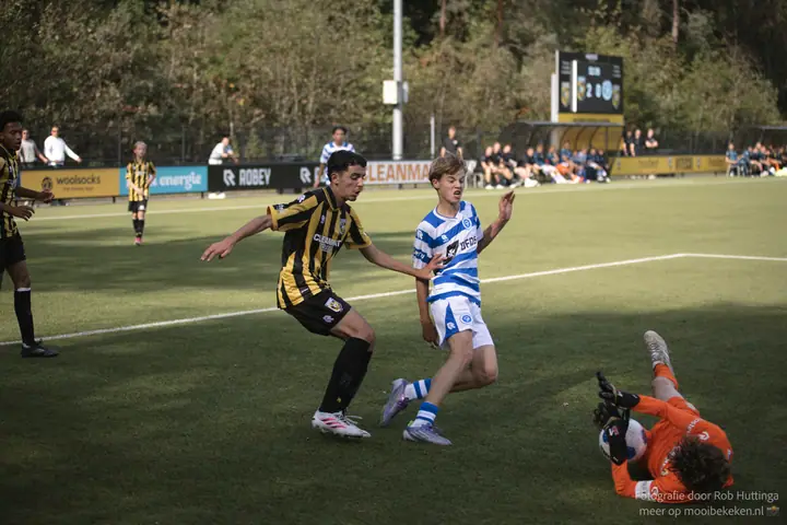 Keeper in oranje duikt op de bal terwijl een speler van De Graafschap en een Vitesse-speler aankomen