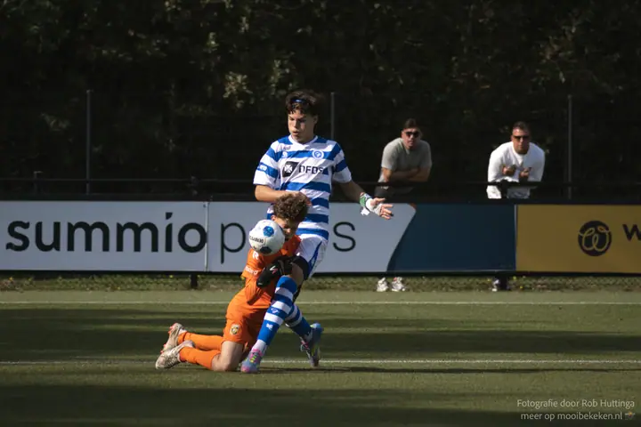 Keeper in oranje klemt de bal tegen zijn borst terwijl een speler van De Graafschap over hem heen stapt