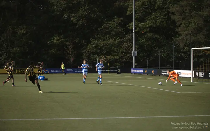 Keeper duikt naar de bal in laag strijklicht, de spelers verspreid over het veld