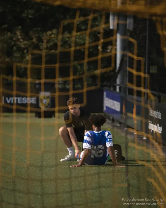 Door het gele doelnet gezien zit een verzorger gehurkt naast nummer 16 van De Graafschap