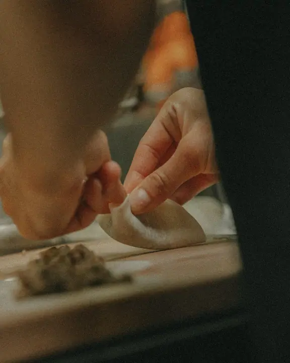 Handen vouwen een dumpling op een houten plank bij Jadda, met de vulling vooraan op tafel