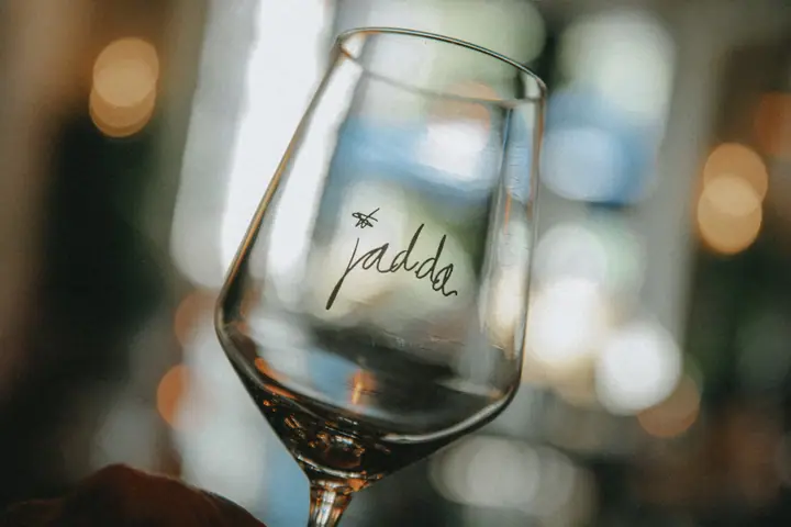 Leeg wijnglas met Jadda-logo omhooggehouden tegen de warme lichtjes van het restaurant