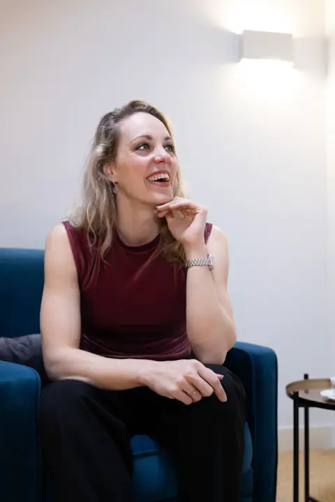 Vrouw in bordeauxrood topje zit lachend in een blauwe fauteuil met haar hand onder haar kin
